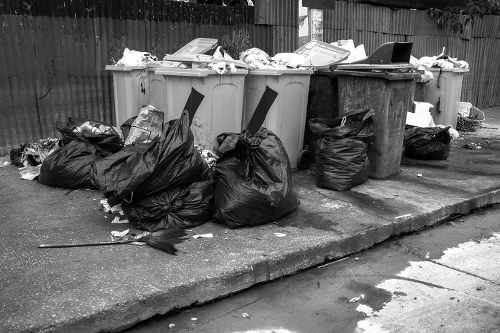A row of large outdoor rubbish bins in a driveway or alleyway, with their lids open and filled with various waste items. Several overflowing black rubbish bags are piled on the ground next to the bins, containing discarded materials. Some trash is spilling out of the bags and scattered on the concrete surface. The bins appear weathered, with different colours, including light grey and darker tones. A wooden fence and some greenery are visible in the background, suggesting a residential area. The scene is captured during daylight, with natural lighting highlighting the texture of the waste and the rough surface of the concrete. This image exemplifies the need for professional rubbish removal services, such as those offered by Garden Clearance Blackheath, to maintain clean and tidy outdoor spaces in the Blackheath area near London.