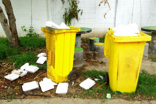 Two large yellow plastic wheelie bins positioned on a paved area beside a white brick wall, with one bin slightly behind the other. The lids of both bins are open, revealing white rubbish inside. Surrounding the bins, there are multiple scattered white disposable food containers and some plastic utensils on the ground, indicating recent rubbish disposal. The area has patches of grass and small plants growing near the wall, and a tree trunk is visible on the left side of the image. Behind the bins, a few small cylindrical concrete stools with coloured tops are arranged on the pavement, contributing to the outdoor setting. The scene appears to be a garden or yard space in Blackheath, suggesting a typical rubbish disposal situation that may benefit from professional waste removal services such as those offered by Garden Clearance Blackheath.