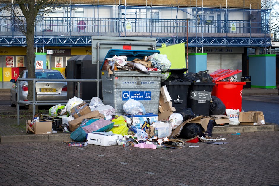 A side view of a white waste collection truck parked on a narrow cobblestone street in an urban area, with a worker dressed in a blue uniform and orange high-visibility vest emptying a blue wheeled tr