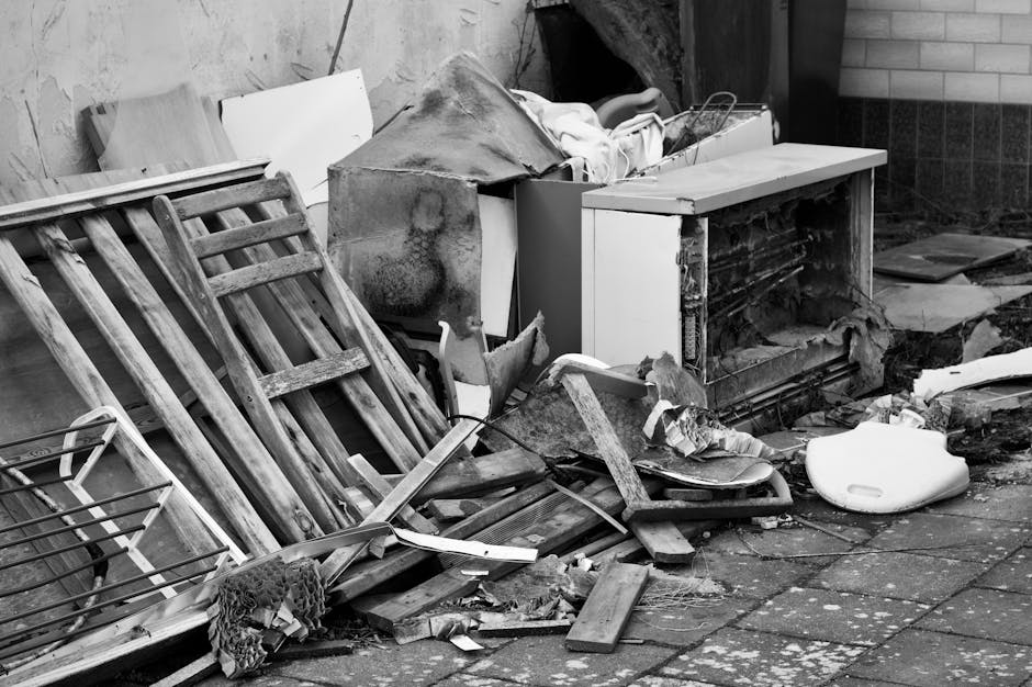 A collection of discarded cardboard boxes, some flattened and others partially collapsed, with visible shipping labels and barcodes, are piled haphazardly on the ground against a rough brick wall and 