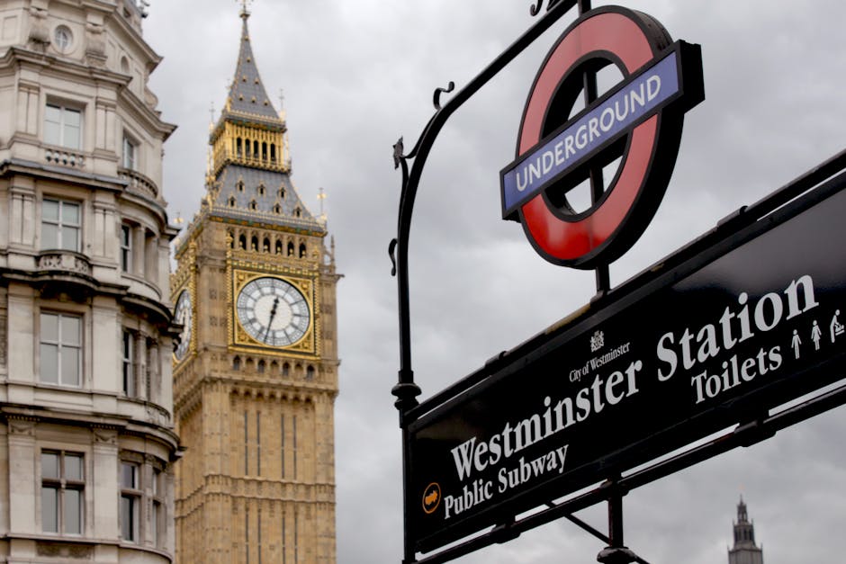 An outdoor view featuring the iconic Big Ben clock tower positioned behind a black and red London Underground sign with a white and black circle, prominently displaying the word 'UNDERGROUND.' Below t